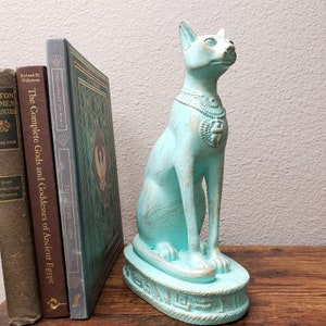 May include: A turquoise blue Egyptian cat statue with a gold scarab beetle on its chest. The statue is sitting on a base with hieroglyphics.
