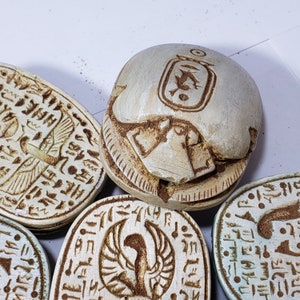 May include: A collection of five white ceramic scarab beetles with intricate carvings and hieroglyphics. The beetles are decorated with brown and gold accents.