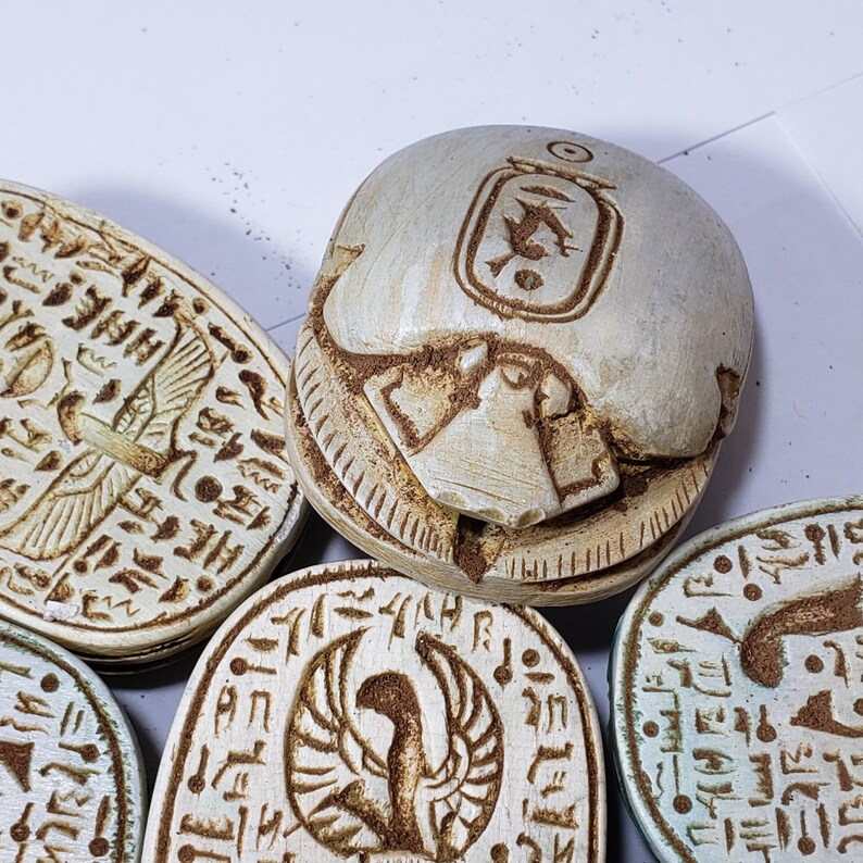 May include: A collection of five white ceramic scarab beetles with intricate carvings and hieroglyphics. The beetles are decorated with brown and gold accents.