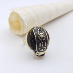 May include: A gold-toned ring with a black oval face featuring hieroglyphic symbols. The ring has a rounded shape with vertical black stripes. A rolled papyrus scroll is in the background.