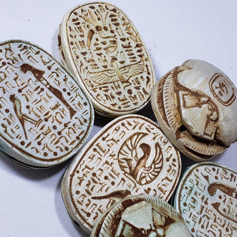 May include: Six ancient Egyptian scarab amulets with hieroglyphic carvings. The amulets are made of a light beige stone and have a rounded, oval shape. The carvings are intricate and detailed, depicting various Egyptian gods and symbols.