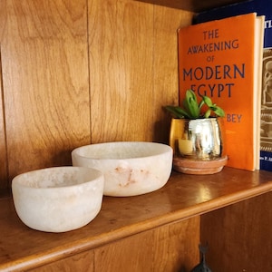 May include: Two off-white, oval-shaped stone bowls sit on a wooden shelf. The bowls have a smooth, polished appearance with subtle variations in color. A small potted plant in a gold-colored pot and a book titled "The Awakening of Modern Egypt" are also on the shelf.