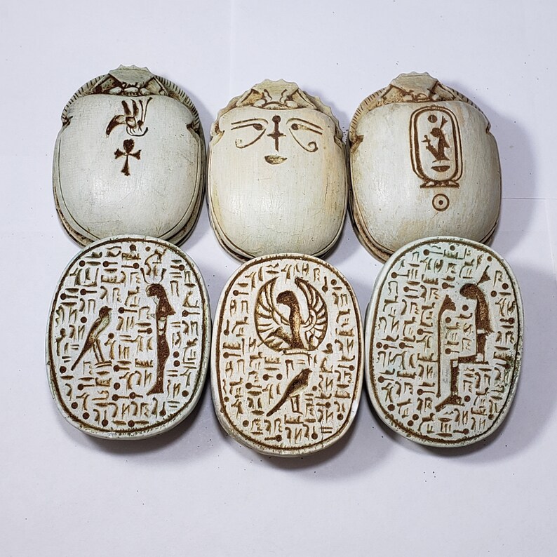 May include: Six white scarab beetle amulets with intricate hieroglyphic carvings. The amulets are oval-shaped and have a raised design. The carvings include images of birds, a winged sun disk, and other Egyptian symbols.