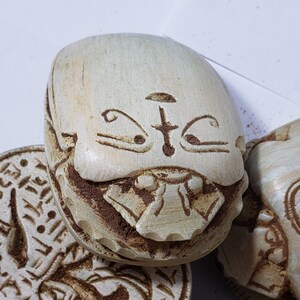 May include: A white ceramic scarab beetle with intricate carvings and a stylized face. The beetle has a detailed design with eyes, a nose, and a mouth. The surface of the beetle is textured and has a slightly worn appearance.