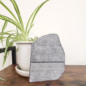 May include: A replica of the Rosetta Stone, a dark gray stone tablet with inscriptions in multiple scripts. The stone is displayed on a wooden surface, with a white potted plant in the background.