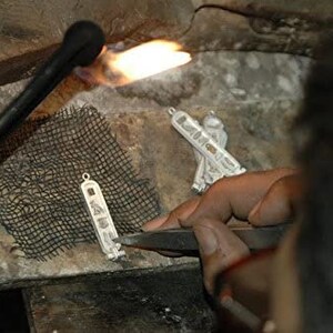 May include: A person uses a torch to heat a silver pendant with intricate details. The pendant is resting on a black mesh surface.