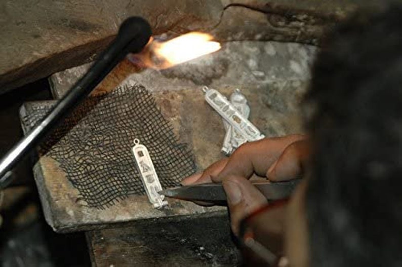 May include: A person uses a torch to heat a silver pendant with intricate details. The pendant is resting on a black mesh surface.