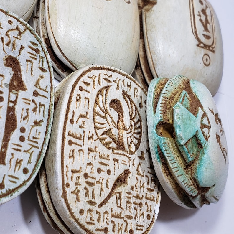 May include: Close-up of six white ceramic scarab beetles with intricate hieroglyphic designs. The scarabs are carved with images of winged creatures and other symbols.