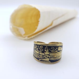May include: A brass-toned ring with a black enamel band featuring a gold-colored engraved design. The ring is set against a white background with a blurred, cream-colored object in the background. The ring's design appears to be ancient.