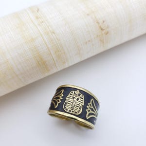 May include: A black and gold cuff bracelet with an Egyptian pharaoh design. The bracelet features a black band with gold detailing of a pharaoh's face and wings. The bracelet is approximately 1 inch wide.