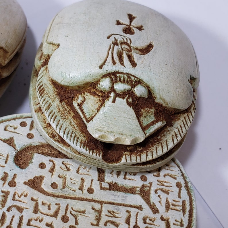 May include: A set of three white ceramic scarab beetles with hieroglyphic markings. The top beetle has a cross-shaped design on its back.
