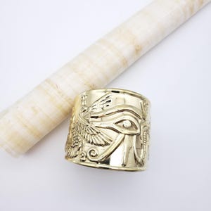 May include: Gold-colored cuff bracelet with an Egyptian Eye of Horus design. The bracelet features intricate embossed details of the eye, wings, and other hieroglyphic symbols. A rolled papyrus scroll is in the background.