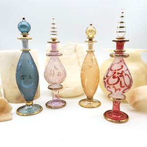 May include: Four colorful glass perfume bottles with gold accents. The bottles are in shades of blue, pink, and peach, with intricate designs and decorative stoppers. The bottles are displayed on a white surface.