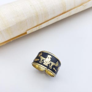 May include: A wide, adjustable ring with a black band and gold-colored accents. The band features a gold-colored bird design. The ring is set against a white background with a papyrus scroll.