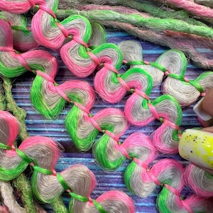 Könnte beinhalten: Nahaufnahme einer Zopf mit rosa, grünen und grauen Haarverlängerungen. Das Haar ist in einem Herzmuster geflochten.