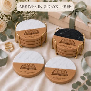 May include: Round coasters featuring a split design of white or black marble and light wood. Each coaster is engraved with an open book icon and the text One More Chapter 2026. Two gold metal wire holders contain stacks of these coasters.