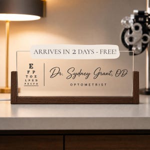 May include: A rectangular clear acrylic desk sign held in a dark wood base. The sign features a black printed eye chart on the left, the name Dr. Sydney Grant, OD in script, and the word OPTOMETRIST in block letters below the name.