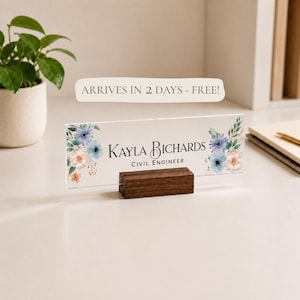 May include: A personalized desk nameplate with a floral design featuring blue and peach flowers. The nameplate is made of clear acrylic and sits in a dark wooden holder. The name "Kayla Bichards" and the title "Civil Engineer" are printed on the nameplate.