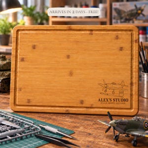 May include: A rectangular bamboo cutting board with a juice groove along the perimeter. The board features a laser-engraved graphic of a propeller airplane, a paintbrush, and a paint pot, with the text ALEX'S STUDIO SCALE MODEL BUILDER.