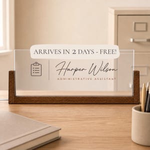 May include: A rectangular desk nameplate featuring a clear acrylic panel held by a dark wood base. The panel displays a clipboard icon, the name Harper Wilson in script, and the title ADMINISTRATIVE ASSISTANT in a clean, sans-serif font.