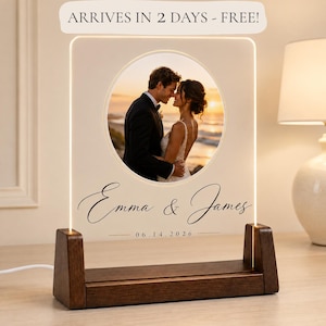 May include: A rectangular acrylic plaque with a circular photo of a couple embracing at sunset, set in a dark wooden base. The plaque features the names Emma & James in black script and the date 06.14.2026 printed below the circular image.