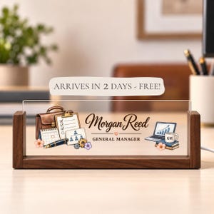 May include: A rectangular clear acrylic desk sign set in a dark wood base. The sign features the name Morgan Reed, the title General Manager, and illustrations of a briefcase, clipboard, calendar, pen, laptop, and coffee mug with GM text.