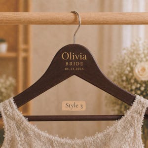 May include: A dark brown wooden clothes hanger with a silver metal hook. The hanger features gold text that reads Olivia, BRIDE, and 06.14.2026. A beige lace garment is draped over the hanger, and a small beige label displays the text Style 3.