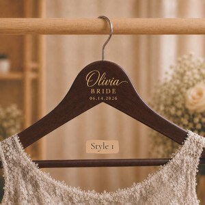 May include: A dark brown wooden clothes hanger with a silver metal hook. The hanger features gold text that reads Olivia BRIDE 06.14.2026 and a rectangular label that reads Style 1. A cream-colored lace garment is draped over the bottom bar.