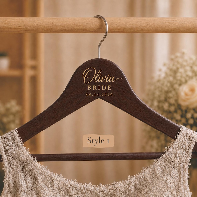 May include: A dark brown wooden clothes hanger with a silver metal hook. The hanger features gold text that reads Olivia BRIDE 06.14.2026 and a rectangular label that reads Style 1. A cream-colored lace garment is draped over the bottom bar.