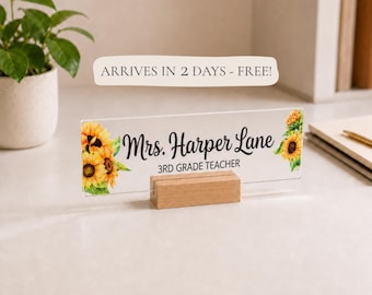 May include: A rectangular clear acrylic desk sign held by a light wood base. The sign features printed yellow sunflower illustrations on the left and right sides, with the black text Mrs. Harper Lane and 3RD GRADE TEACHER centered on the front.