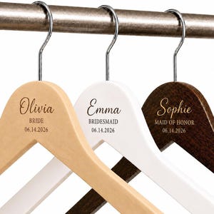 May include: Three wooden clothes hangers with silver metal hooks, featuring engraved names and titles. The first is natural wood with Olivia, BRIDE, 06.14.2026. The second is white with Emma, BRIDESMAID, 06.14.2026. The third is dark wood with Sophie, MAID OF HONOR, 06.14.2026.