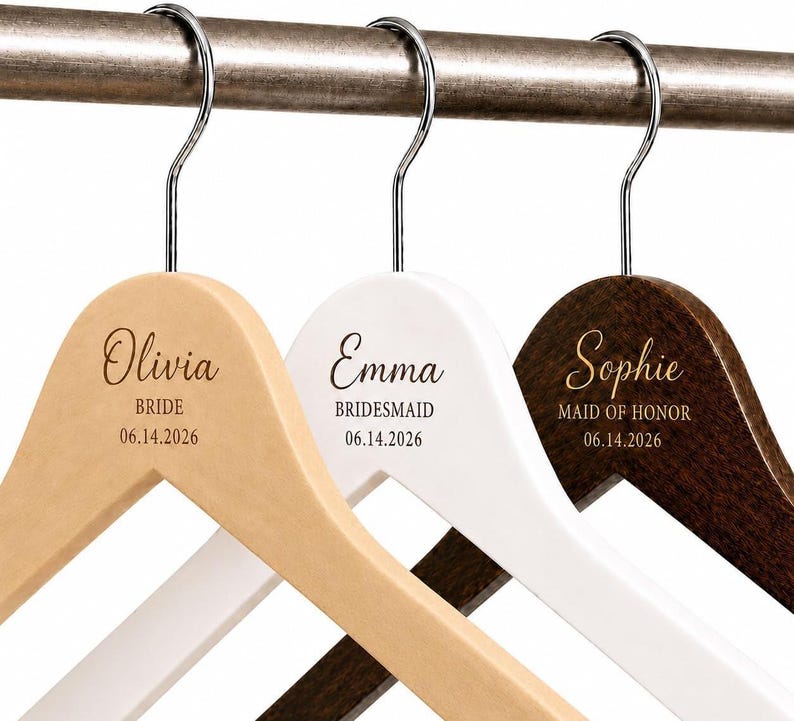 May include: Three wooden clothes hangers with silver metal hooks, featuring engraved names and titles. The first is natural wood with Olivia, BRIDE, 06.14.2026. The second is white with Emma, BRIDESMAID, 06.14.2026. The third is dark wood with Sophie, MAID OF HONOR, 06.14.2026.