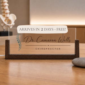 May include: A rectangular clear acrylic desk sign held by a dark wood base. The sign features a teal spinal column graphic, the text Dr. Cameron Wells, and the word CHIROPRACTOR in black lettering. The sign is positioned on a wooden surface.