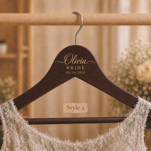 May include: A dark brown wooden clothes hanger with a silver metal hook. The hanger features gold text that reads Olivia, BRIDE, and 06.14.2026. A beige rectangular label with the text Style 2 is centered on the hanger above a cream lace garment.