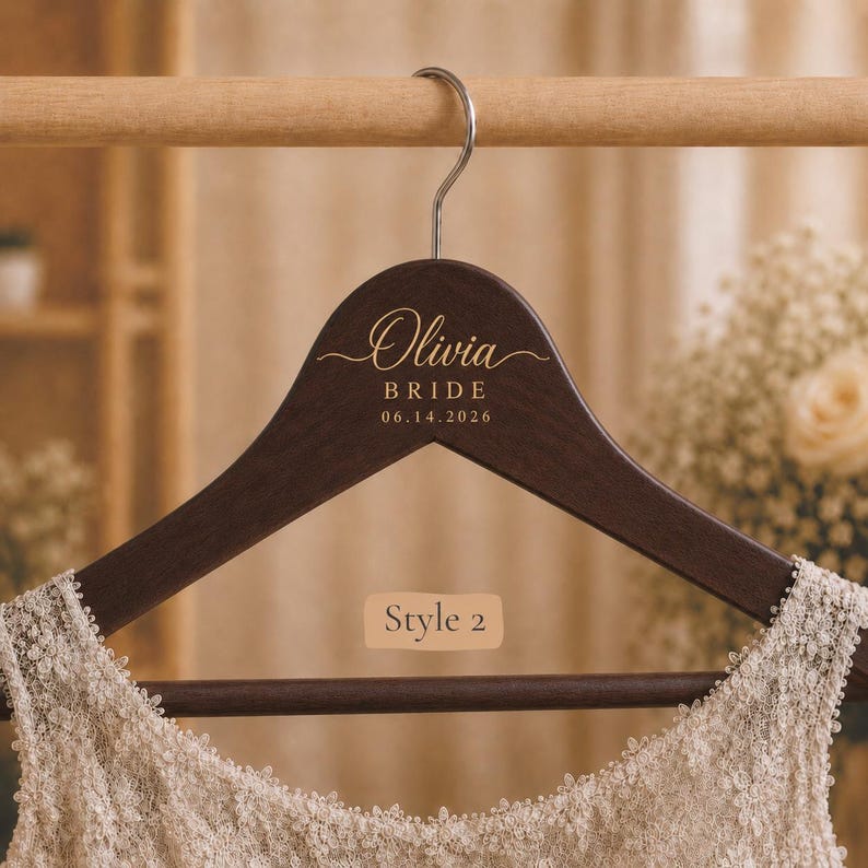 May include: A dark brown wooden clothes hanger with a silver metal hook. The hanger features gold text that reads Olivia, BRIDE, and 06.14.2026. A beige rectangular label with the text Style 2 is centered on the hanger above a cream lace garment.