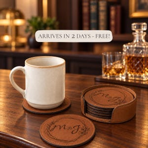 May include: A set of round brown leather coasters featuring a laser-engraved floral wreath design with the letters M&J in the center. One coaster sits under a cream-colored ceramic mug, while others are stacked inside a matching leather holder.