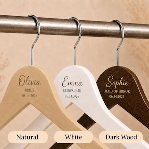 May include: Three wooden clothes hangers with silver metal hooks, featuring custom engravings. The natural wood hanger says Olivia BRIDE 06.14.2026, the white hanger says Emma BRIDESMAID 06.14.2026, and the dark wood hanger says Sophie MAID OF HONOR 06.14.2026.