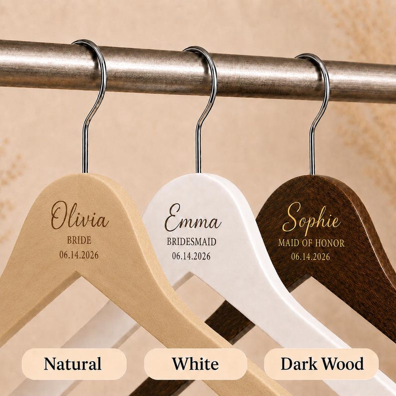 May include: Three wooden clothes hangers with silver metal hooks, featuring custom engravings. The natural wood hanger says Olivia BRIDE 06.14.2026, the white hanger says Emma BRIDESMAID 06.14.2026, and the dark wood hanger says Sophie MAID OF HONOR 06.14.2026.