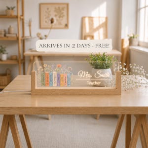 May include: A rectangular light-up acrylic sign set in a light wood base. The acrylic features a colorful illustration of a row of books with floral accents, the text "Mrs. Smith" and "Teacher," and individual book spines labeled with words.