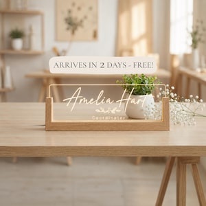 May include: A lighted desk nameplate with the name "Amelia Hart" in elegant script. The nameplate is set in a wooden frame and sits on a wooden table. A small potted plant and flowers are next to the nameplate.