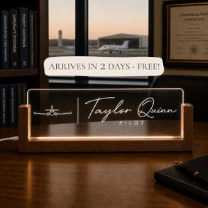 May include: A rectangular clear acrylic desk sign with a wooden base, featuring an illuminated white line drawing of an airplane, the name Taylor Quinn, and the word PILOT in a script and sans-serif font combination.