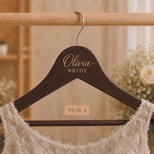 May include: A dark brown wooden clothes hanger with a silver metal hook. The hanger features the gold text Olivia and BRIDE in a script and sans-serif font. A beige rectangular label with the text Style 4 is visible below the hanger.