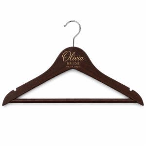May include: A dark brown wooden clothes hanger with a silver metal hook. The center of the hanger features gold engraved text that reads Olivia, BRIDE, and the date 06.14.2026. The hanger includes notches on both ends and a horizontal bottom bar.