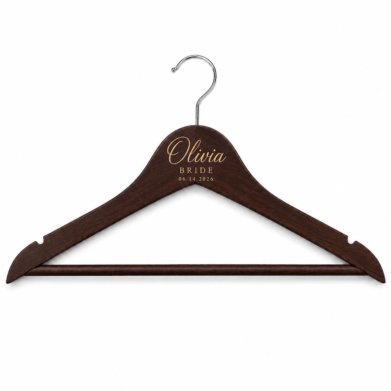 May include: A dark brown wooden clothes hanger with a silver metal hook. The center of the hanger features gold engraved text that reads Olivia, BRIDE, and the date 06.14.2026. The hanger includes notches on both ends and a horizontal bottom bar.