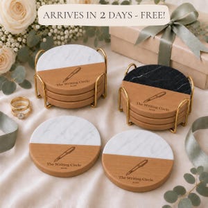 May include: Round coasters featuring a split design of white or black marble and light wood. Each coaster displays an engraved fountain pen icon with the text The Writing Circle 2026. Two sets are held in gold metal wire stands on a fabric surface.