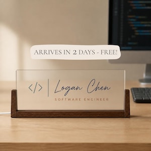 May include: A rectangular frosted acrylic desk sign held by a dark brown wooden base. The sign features a code bracket symbol, the name Logan Chen in a dark blue script font, and the text SOFTWARE ENGINEER in a brown sans-serif typeface.