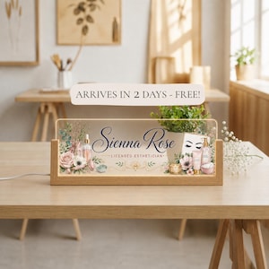 May include: A rectangular acrylic sign with a wooden base featuring the text Sienna Rose Licensed Esthetician, decorated with illustrations of pink roses, eucalyptus leaves, skincare bottles, a jade roller, and a small framed eye graphic.