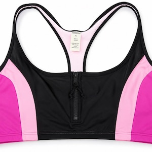 May include: A black racerback sports bra featuring a front zipper closure, light pink side panels, and vibrant magenta outer sections. The interior label displays size XL, fabric composition, and origin details in clear black text on a white background.