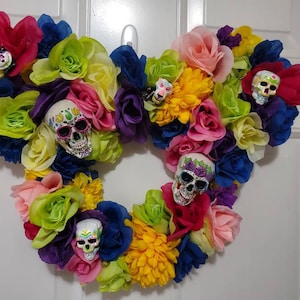 May include: A heart-shaped wreath decorated with colorful artificial flowers and sugar skull ornaments. The wreath is made of a variety of flowers in shades of pink, red, yellow, green, blue, and purple.