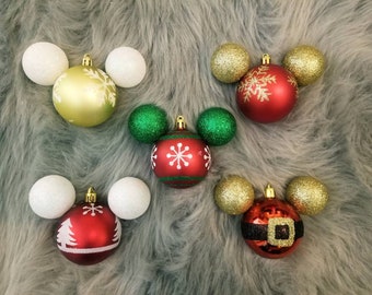 Puede incluir: Cinco adornos navideños con forma de cabeza de Mickey Mouse. Los adornos son rojos, verdes, dorados y blancos con diseños de copos de nieve y otras fiestas.